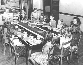 11 6x8 grade white children sitting at a dinning room table the teacher is at the head of the table there might be a schooll cafiteria behind. All the plates are empty but every plate setting would be 4H contest level today there are two american flags as center pieces