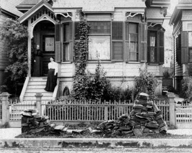 Image of a partly depleted slab wood pile in front of a home along the sidewalk in late 1890s. Women is standing on the stairs leading into her home.