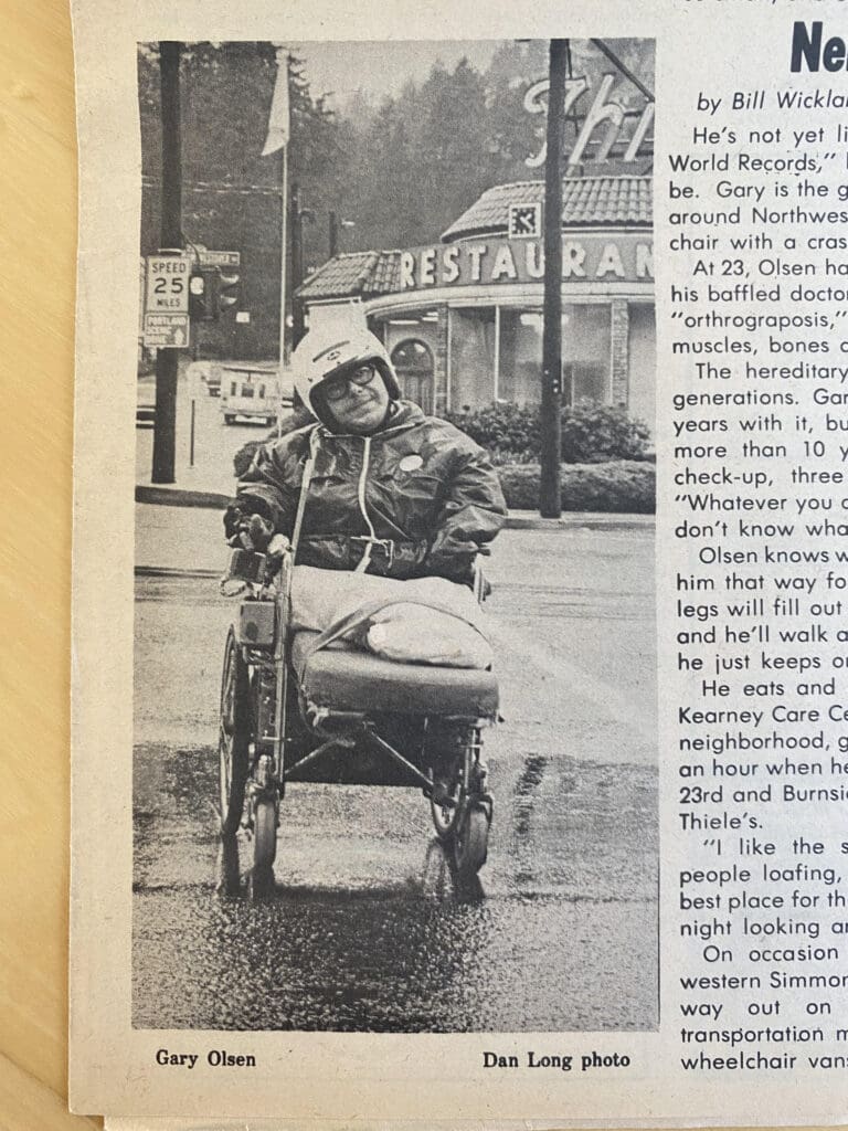 John in his wheel chair in the rain. Dan Long photo from an issue of the Neighbor. Back Ground is the long demolished Henry Thile's Restaurant. The clock on the restaurant reads 1:25pm. John is wearing a helmet and a water proof jacket. He has on glasses