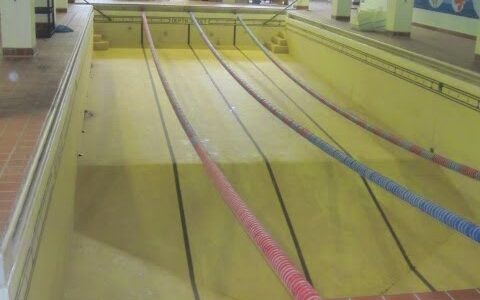 Image of the pool in the basement of MLC School. The water has been drained out but the lane dividers are still attached hanging like party decorations. Part of a mural of seas creature is on the right. This is taken from the deep end of the dry pool where there was once a diving board.