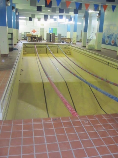 Image of the pool in the basement of MLC School. The water has been drained out but the lane dividers are still attached hanging like party decorations. Part of a mural of seas creature is on the right. This is taken from the deep end of the dry pool where there was once a diving board.