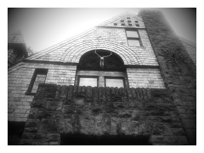 Black and White exterior image of a stone house with a stage head under an archway. A very unusual example of Queen Shingle Style because the shingles are made of slate. The chimney is massive with decorative metal sun on the basalt rock that starts at the basement level and then contrasts with the shingles..