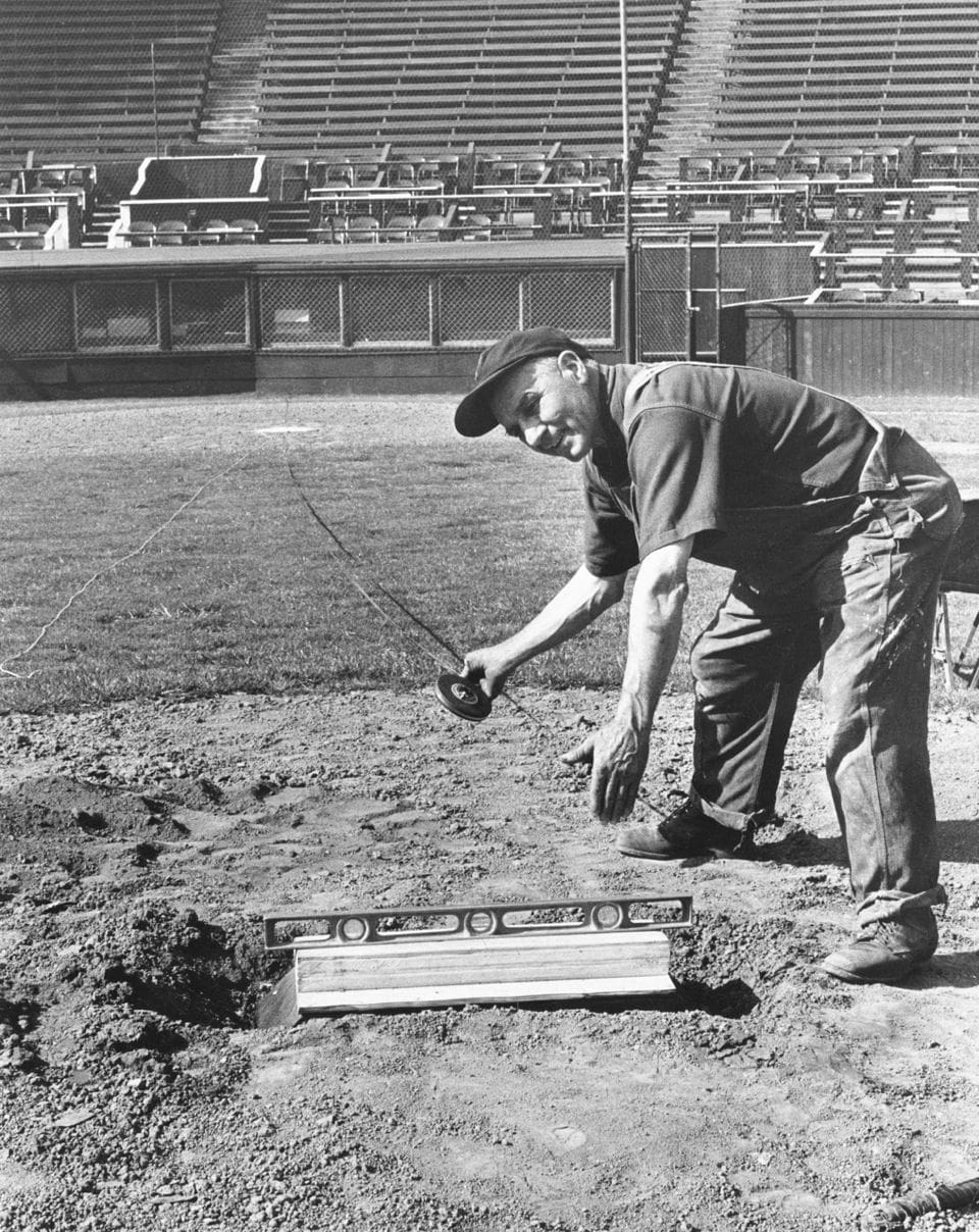 Rocky the ground keeper at Vaughn Street Ballpark holding a level near hoe plate leaning over -grass turf perfectionist