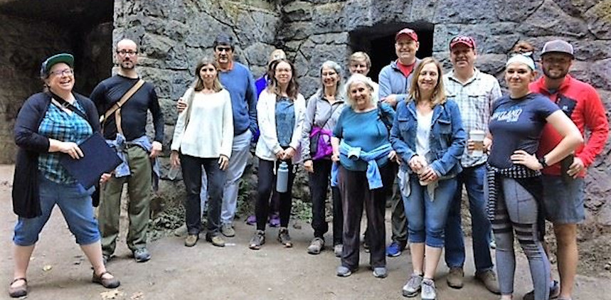 Tanya March owner on Left. 13 tour guests posing in Forest Park in front of a 1929 comfort station. Nicknamed the Witch's house, the basalt stone looks too fancy to be a restroom.