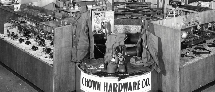 Display cases inside Chown Hardware Co. There are guns under the glass in the case on the right. Possibly fishing reels or maybe door knobs in the case on the left. Display of riffles in the background. In the foreground boot and rain gear.