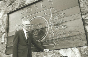 Text: wooden sign is engraved Max is blocking some of the sign. There are rose retails and the word at top Roses at bottom cocktails.