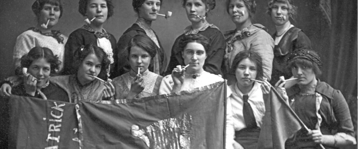 A dozen women with tobacco pipes in their mouths holding up a banner that says St Patrick and has a harp on it. Very silly poses but not a candid image.