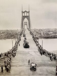 St Johns Bridge with cars a floats covered with flowers driving in one lane people standing on both sides of suspension bridge.