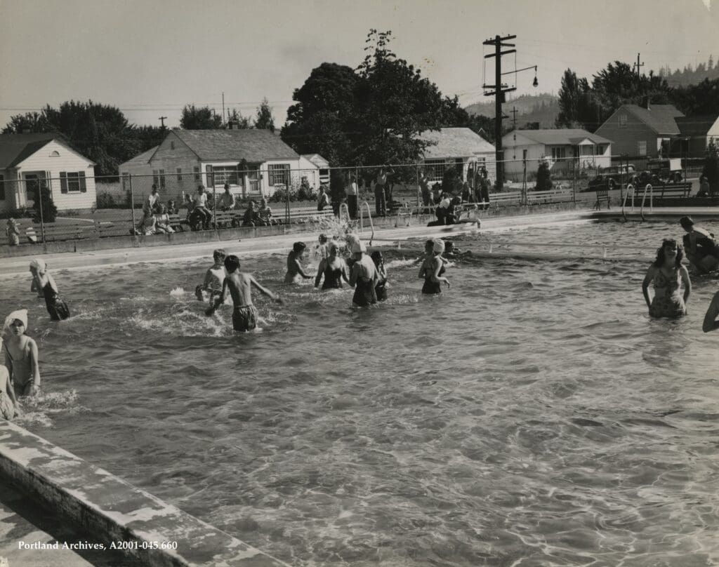 a open air pool suburban style home past a chain link fence in the background kids playing in a pool