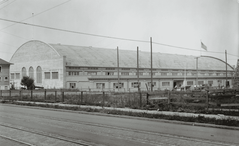 Wide angle black and white image of a super block long building that has a roof like a quonset hut and and entry.