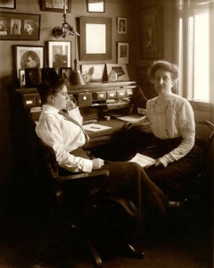 Two women with pompadores in an office. Many images of 1900s stars head shots on the wall above a roll down desk.