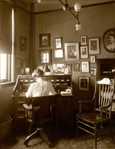 Photograph of Lois Steers seated at a desk in a corner of the Steers-Coman office. Her back to the camera, she is working at the desk, wearing a shirtwaist blouse and with her hair done in the pompadour style popular in the early years of the 1900's. A second chair, empty, sits near the desk. The walls visible in the photograph are covered with portraits of musical celebrities. There are also framed portraits sitting on the top of the desk.