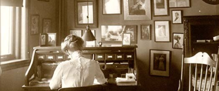 Photograph of Lois Steers seated at a desk in a corner of the Steers-Coman office. Her back to the camera, she is working at the desk, wearing a shirtwaist blouse and with her hair done in the pompadour style popular in the early years of the 1900's. A second chair, empty, sits near the desk. The walls visible in the photograph are covered with portraits of musical celebrities. There are also framed portraits sitting on the top of the desk.