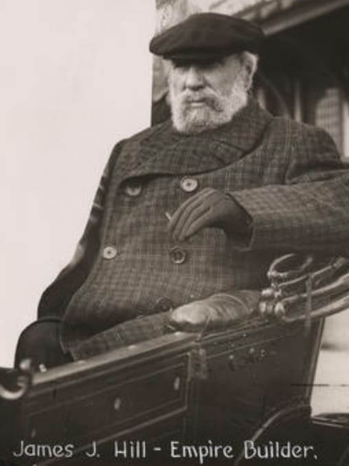 Man sitting in the back seat of a car as if he is sitting on a throne text on Image James J. Hill - Empire Builder there is a building framing the shot might be a train station. Plaid coat gloves might have a pen or a cigarette in his hand. A cap is on his head he is old enough to have thinning hair to hide.