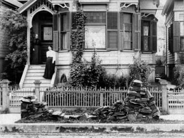 a depleted pile of slabwood a woman poses far away from the photografer leaning up against rail of a charming vernacular wooden home
