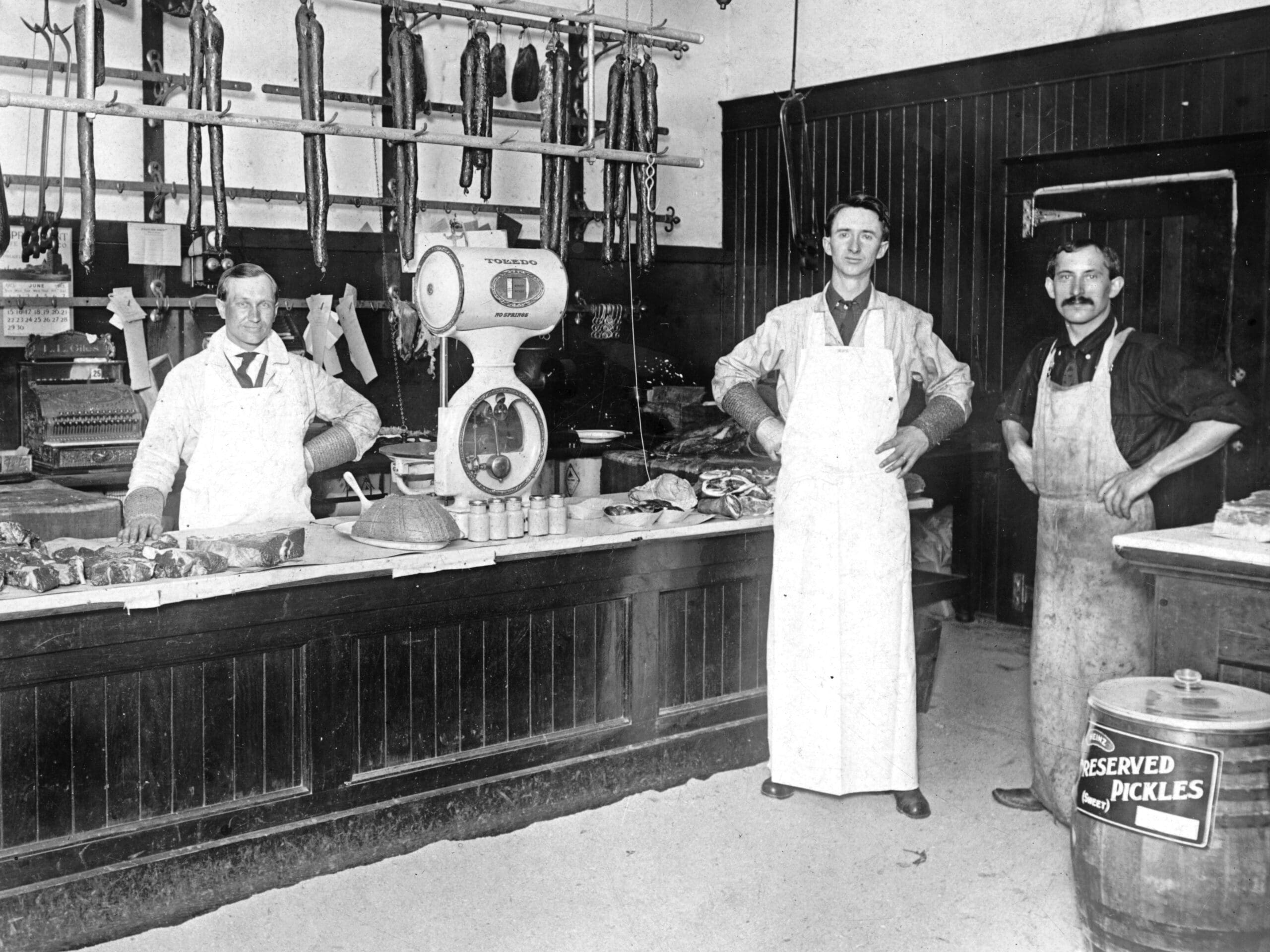 Two men behind a butcher block counter, two additional men on the right. A huge wooden barrel of pickles in the for ground. There are items of meat handing from the ceiling in the back ground. There is a layer of sawdust on the floor.