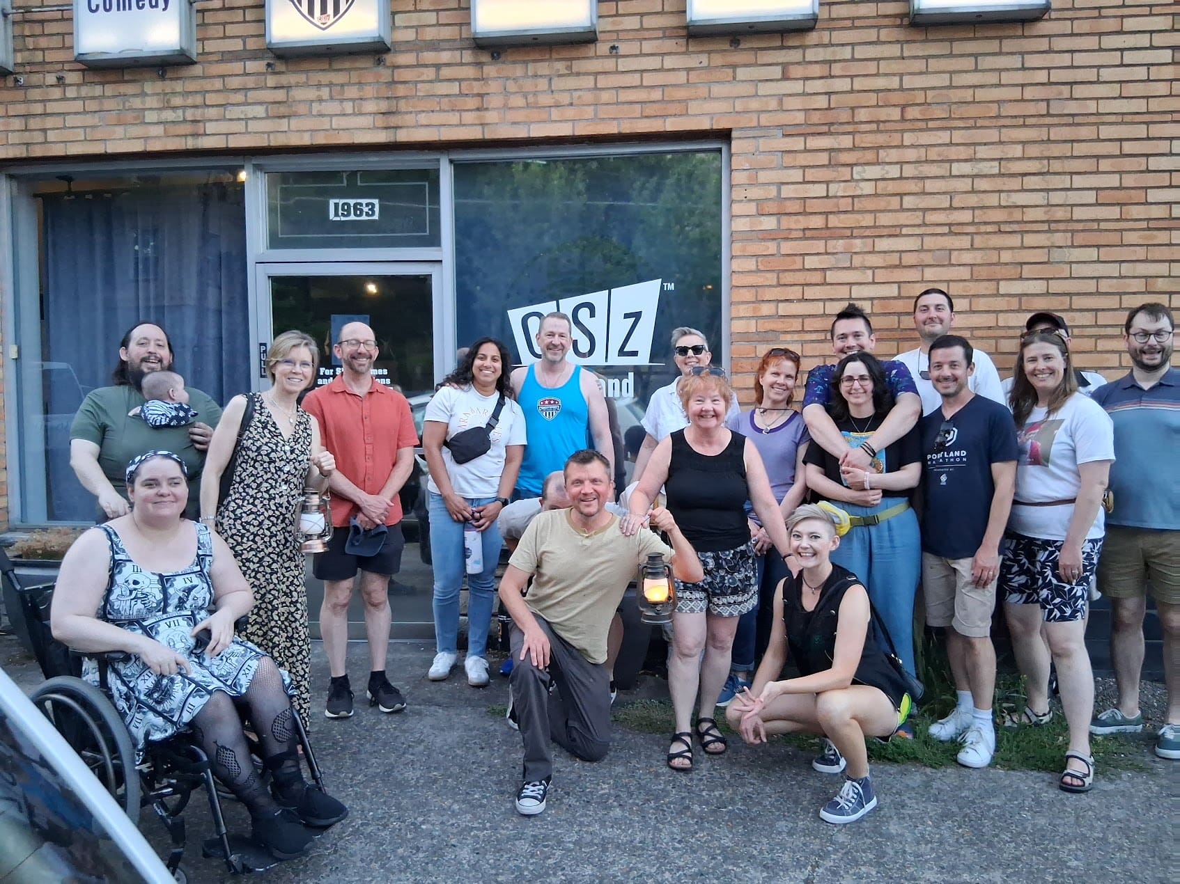 Woman in wheel chair along wit a group of 15 friends and staff posing in front of the Comedy Club. A single story masonry building