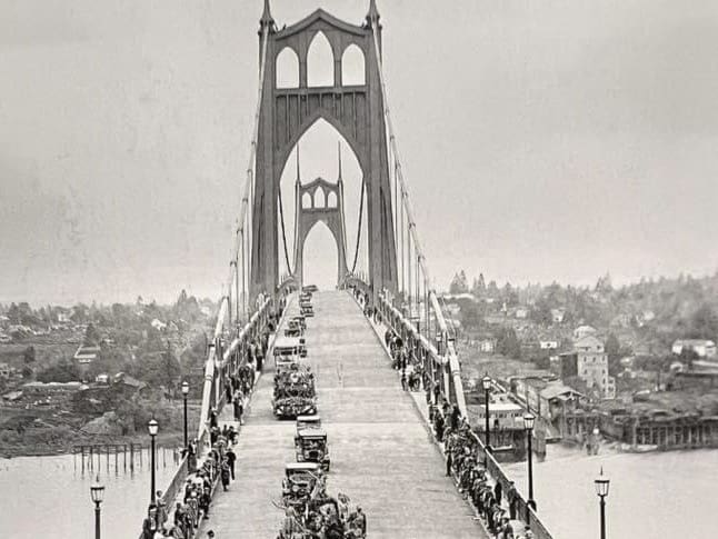 Image of the St. Johns bridge in black and white taken during the Rose Festival Parade. Skyline of St. Johns in the distance. This image is focused on cars driving on the bridge deck in a single line heading west.