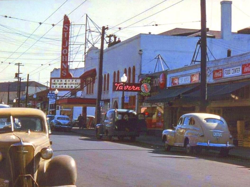 This is a rare color street view of Lombard in St. Johns looking east with The St. Johns theater at the far end of the shot on the right side of street view you only see the south side of the street as far as buildings a car windshield and fender is on the left no lines on the street and no potholes also street trees are not present not that its winder but if they are there they are babies
