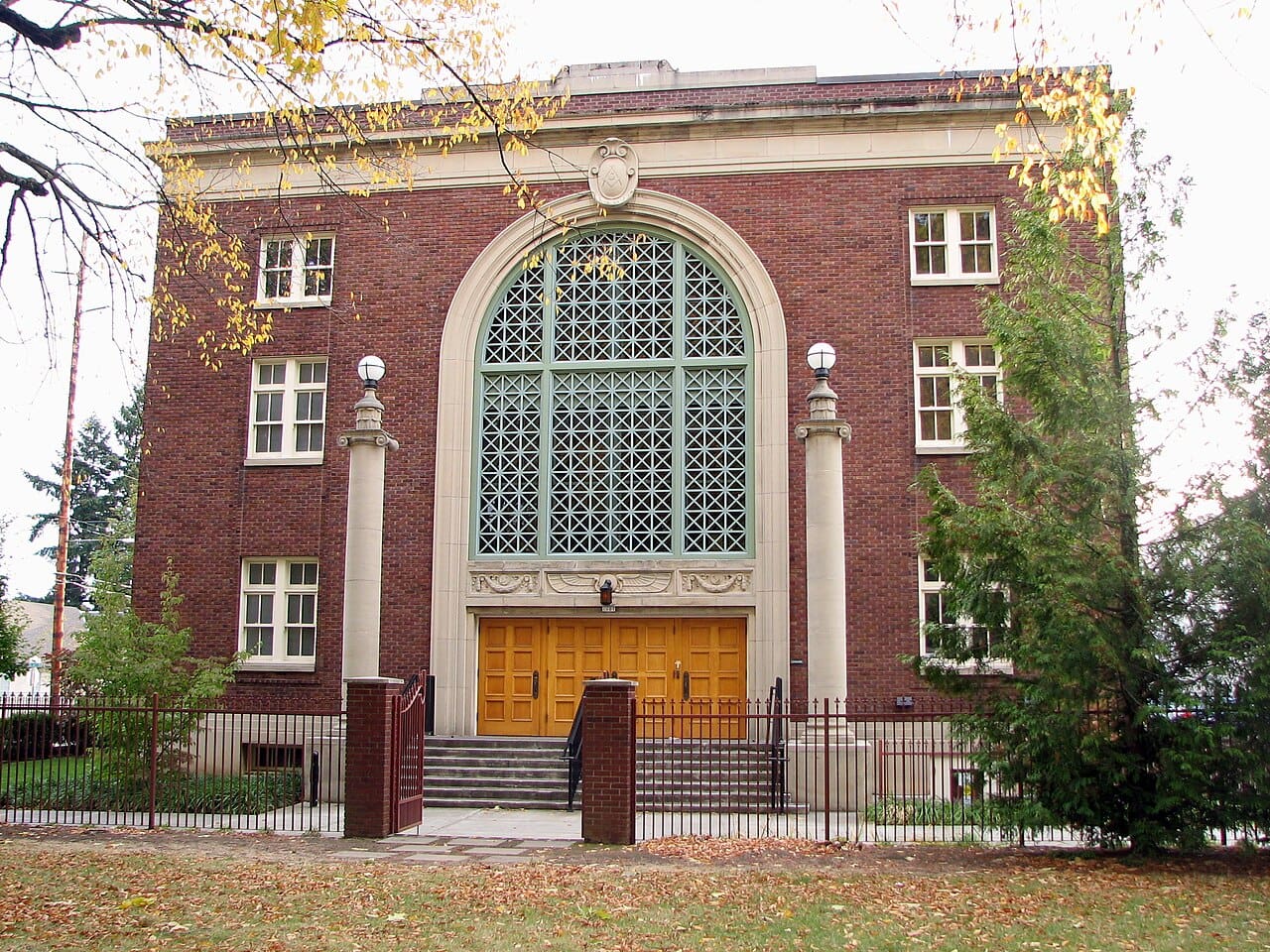 This is a daylight image of a three story brick building with a huge three story arch with a first story bank of doors that are reached by steps. There are two columns with round balls on top closer to the viewer than the building one on each side of the doors.