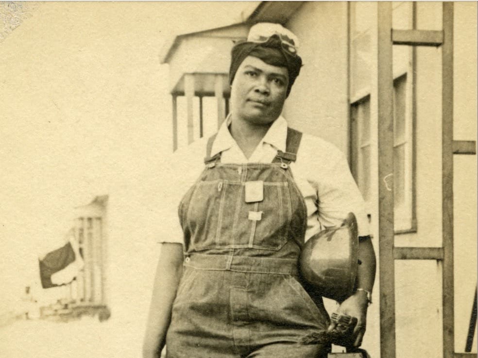 Cropped Image of a Black women in overalls hair is covered in a scarf she might be holding a safety helmet under her arm