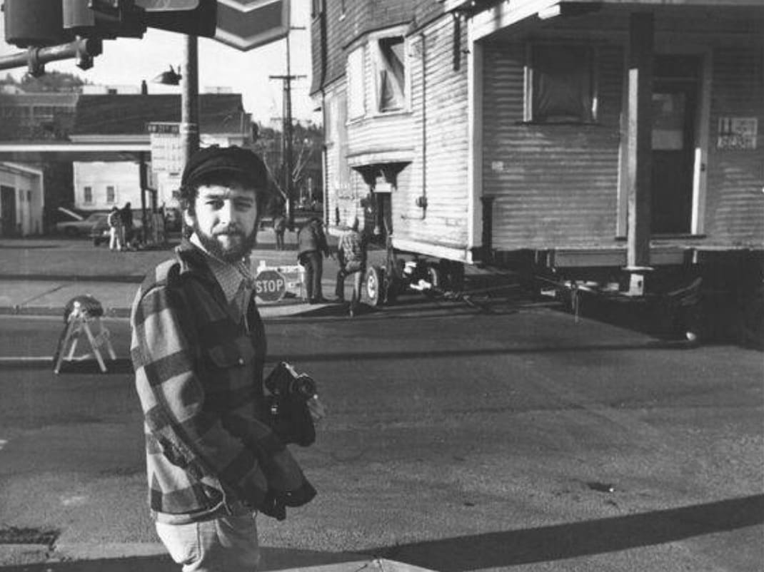 Mike Ryerson is in his 40s he has a camera in hand and behind him is a building being rolled on a street to save it friom demolition