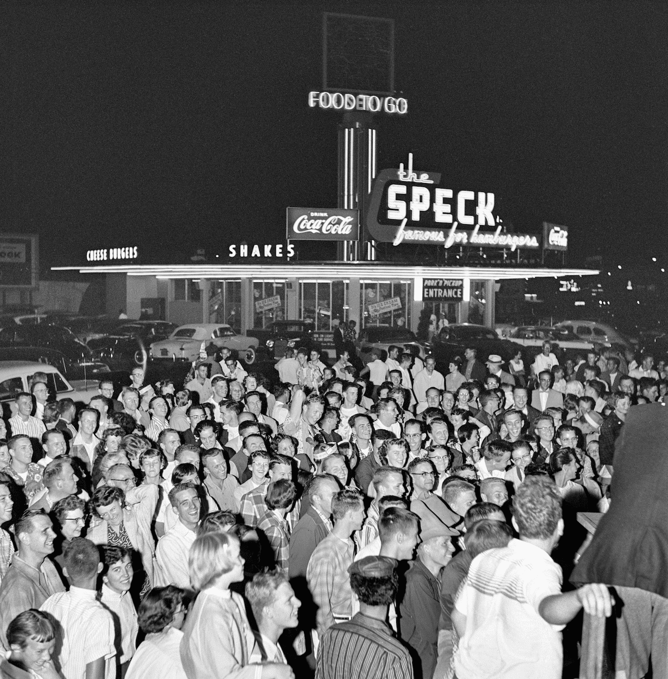 The small drive in dinner theSPEACK in neon with neon lights run up a tower with neon saying FOOD TO GO, Neao the SPECK famous hamburgers also in neon SHAKES, CocaCola - There is a sing over the door "Entrance". 1950s on Foster Road 100 of people are standing around most are facing to the right.