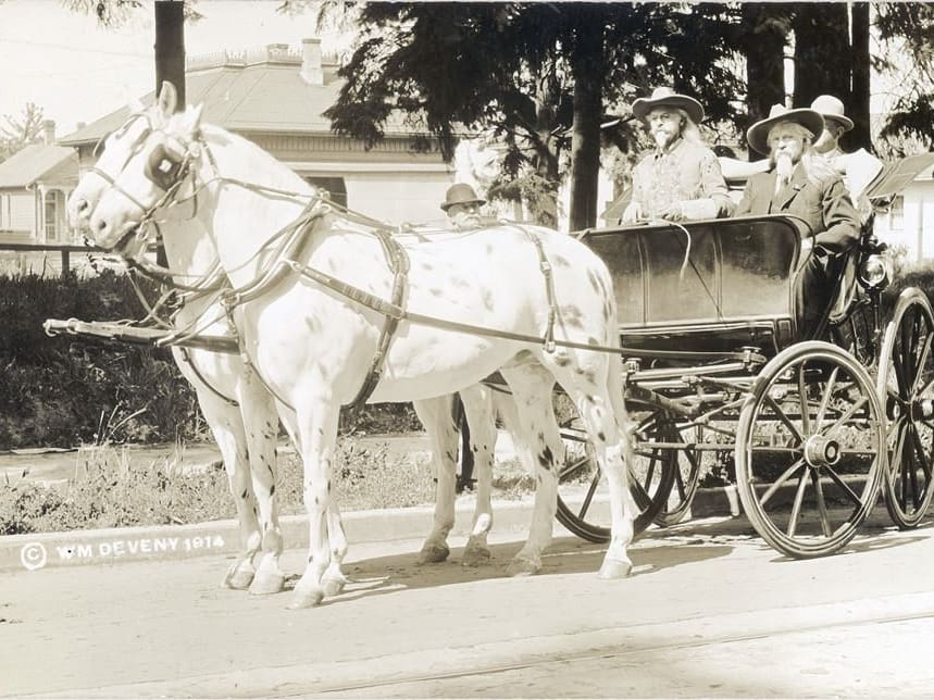 A team of horses pulling a carriage with two look a like "Wild Bills" in the frint you can't see the face of their passaer and there is a man in a bowler hat and a house in the back ground