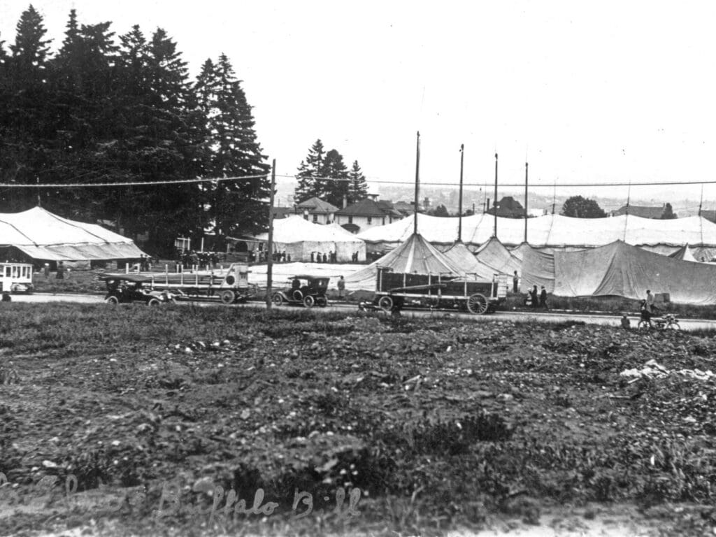 The fore ground is a weedy plot of land on a road are early 1900s vehicles in between the road and homes looking east are circus tents the big top is at a low point the poles stand proud and there are a cluster of trees on the left